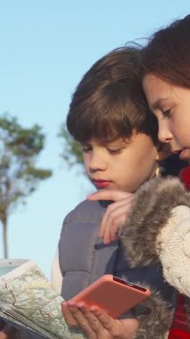 İki maceraperest çocuk, bir oğlan ve bir kız, parlak mavi bir gökyüzünün altında dururken hevesle bir harita çalışıyorlar, ifadeleri merakla dolu. Sıcak bir şekilde giyinen kız, rahat, beyaz kürklü bir ceket ve canlı kırmızı bir eşarp giyiyor.