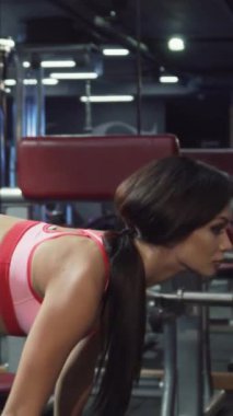 Gösterişli, modern bir spor salonunda, uzun, koyu saçlı, at kuyruklu, kendini adamış genç bir kadın halterle kendinden emin bir şekilde öne eğiliyor. Parlak pembe atletik bluzu ve siyah taytları, güçlü fiziğinin ve enerjisinin altını çiziyor.