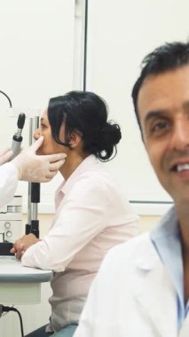 Sıcak kalpli bir doktor ön planda kendine güveni olan, yaklaşımlı bir ışık saçar. Arkasında, kendini işine adamış bir göz doktoru bir kadının gözlerini titizlikle inceliyor. Parlak, modern bir tıp ofisinde besleyici ve profesyonel bir atmosfer yaratıyor.