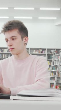 Büyüleyici düşük açılı bir çekim, pembe kazak giymiş, parlak ve modern bir kütüphanede laptopuna odaklanmış genç bir erkek öğrenciyi gözler önüne seriyor. Renkli kitaplıklarla dolu, yüksek kitaplıklarla çevrelenmiş,