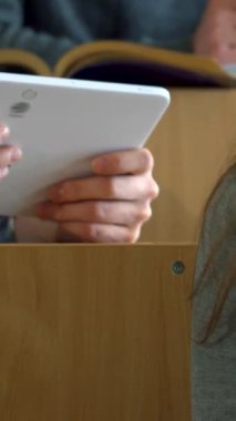 Dinamik bir kolej konferans salonunda, genç beyaz bir erkek öğrenci kadın sınıf arkadaşıyla parlak bir tablet üzerinde büyüleyici sezgilerini paylaşıyor ve işbirliği ruhunu teşvik ediyor. Enerjik atmosfer, odaklanmış ifadelerle zenginleşir.