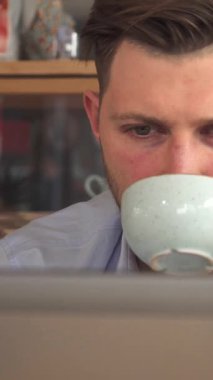 Genç, sakallı bir iş adamı, yoğun bir şekilde odaklanmış, bir kafede özenle dizüstü bilgisayarında yazarken dumanı tüten bir fincan kahvenin tadını çıkarıyor. Sıcak ahşap aksanlar ve yumuşak ortam ışıklarıyla süslenmiş davetkar atmosfer mükemmel bir zemin oluşturur.