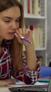 Kendini işine adamış beyaz bir kız öğrenci, dizüstü bilgisayarına dalmış, yüksek kitaplıklarla çevrili kalemini ısırıyor. Titrek damalı gömleği, akademik kararlılığını yansıtan huzurlu kütüphane dekoruyla güzel bir tezat oluşturuyor.