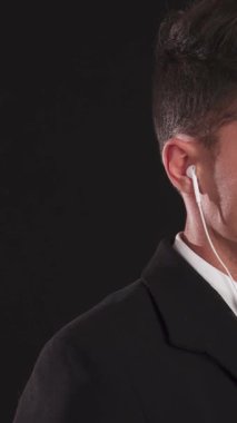 Siyah takım elbiseli çarpıcı bir genç, parlak siyah bir arka plana karşı duruyor, kendine güven ve otorite yayıyor. Odaklanmış bir ifadeyle, kulaklığından dikkatlice dinler. Avucunu keskin bir şekilde uzatır.