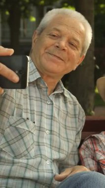 Her ikisi de canlı bir park bankında oturan neşeli torunuyla selfie çekerken hoş bir kıdemli adam neşeyle ışıldıyor. Moda bir şapkası olan küçük çocuk yemyeşil bir zemin ve benekli bir güneş ışığına karşı mutluluk saçıyor.