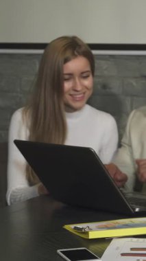 Modern bir ofis ortamında, uzun sarı saçlı odaklanmış genç bir kadın çenesini eline dayayıp, enerjik iş stratejilerini tartışan meslektaşlarını dikkatle izliyor. Önündeki parlak dizüstü bilgisayar ve renkli belgeler dağınık.