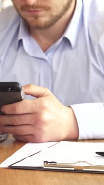 Şık, sarışın bir işadamı, akıllı telefonuna dalmış, kırsal bir ahşap masada elinde bir not panosu ve kalemle oturuyor, ve modern üretkenlik hissi yayıyor. Yumuşak ışıklandırma ve sıcak tonlarla, davet edici kafe ambiyansı, mükemmel bir karışım oluşturur