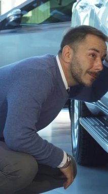 İyi bakımlı sakallı odaklanmış bir adam çarpıcı gümüş bir arabanın yanına çömelmiş, çarkını keskin bir ilgi ve tatmin edici bir gülümsemeyle inceliyor. Yumuşak ışıklandırma ve cilalı yüzeylerle dolu çağdaş sergi salonu lüks atmosferi geliştirir.