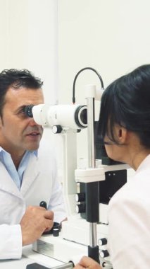 Parlak, modern bir ofiste, iki neşeli doktor bir hastayla sıcak ilişki kurar, biri gözlerini incelemek için uzman bir cihaz kullanır. Davetkar atmosfer dostça sohbetler ve profesyonel ilgiyle dolu.