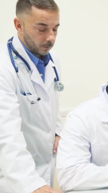 Parlak ve modern bir ofiste, iki dost canlısı erkek doktor bir hastayla karşılaşır, biri gözlerini incelemek için yüksek teknolojili bir cihaz kullanır. Sıcak atmosfer profesyonellik ve şefkat yayarak, sağlık ve refaha olan bağlılıklarını vurguluyor.