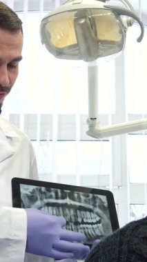 Dost canlısı, beyaz sakallı beyaz önlüklü beyaz tenli beyaz bir dişçi. Zarif bir dokunmatik tablet üzerinde röntgen görüntüsü sergilerken erkek bir hastayla sıcak etkileşime giriyor. Modern bir dişçi kliniğindeki bu davet sahnesi