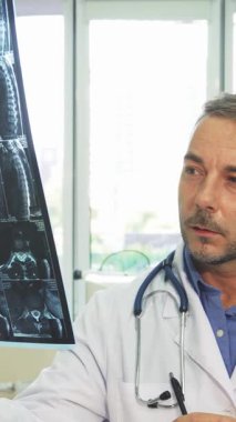 Gıcır gıcır beyaz laboratuvar önlüğü giymiş kendini adamış genç bir adam X-Ray görüntüsünü yüksek tutuyor ve parlak, modern ofisinde yoğun bir şekilde inceliyor. Steteskop boynundaki profesyonel titreşimi artırırken, parlak mobilyalar ve yumuşak bej rengi...