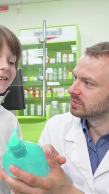 Kendini işine adamış beyaz önlüklü bir erkek eczacı, parlak bir turkuaz şampuan şişesini zevkle inceleyen heyecanlı küçük bir çocukla sıcak etkileşime giriyor. Bu hoş an renkli bir eczanenin canlı atmosferini gözler önüne seriyor.