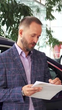 Doğal ışıkla dolu şık, modern bir araba galerisinde, mutlu bir alıcı ve cilalı bir satıcı, araba alımlarını neşeyle sonuçlandırmak için kontratı imzalarken göz alıcı bir gülümseme paylaşıyorlar. Şık ekose ceketli adam coşkuyla bakarken,