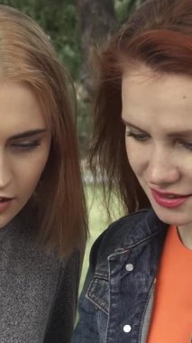 Yeşilliklerle dolu yemyeşil bir parkta, biri parlak kızıl saçlı, diğeri altın sarısı bukleli iki genç kadın, bir akıllı telefonu paylaşıyor, yüzleri kahkaha ve heyecanla aydınlanıyor. Sıcak yaz güneşi onların gündelik güneşlerine neşe saçar.