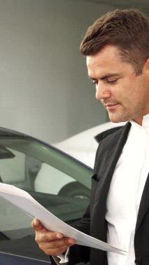 İyi bakımlı sakallı ışıldayan bir adam, çağdaş bir galeride lüks araçlarla çevrili yeni arabasının anahtarlarını gururla sergilerken neşe saçıyor. Sivri siyah bir takım elbise ve şık bir damalı gömlek giymişti. Kendine güveni tamdı.