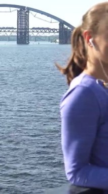 Büyüleyici bir sahnede uzun, koyu renk saçları at kuyruğu olan genç bir kadın parıldayan nehrin kenarındaki korkuluklara yaslanıyor. Canlı bir menekşe bluz giyip, dikkatle kulaklıklarından dinliyor. Bakışları dingin bir odak noktası oluşturuyor.