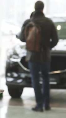 Modern bir araba galerisinde şık sakallı genç bir adam duruyor, akıllı telefonuyla derinden meşgul, parlayan araçların arkaplanına mesajlar yazıyor. Bu dinamik sahne teknoloji ve lüksün kaynaşmasını güzel bir şekilde gösteriyor.