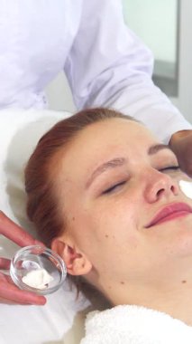 Kendini adamış, el değmemiş beyaz önlüklü bir kozmetik uzmanı müşterisinin yüzüne nazikçe lüks bir krem sürerek doğal güzelliğini arttırıyor. Bir elinde narin bir fırça, diğer elinde kremalı bir kaseyle, güzellik uzmanı...