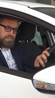 Şık sakallı ve gözlüklü sofistike olgun bir erkek müşteri, bayilikte modern bir arabanın gösterişli iç mekanını keşfetmekle son derece meşgul. Rahatça oturur, direksiyona hafifçe dokunur, heyecan saçar ve...