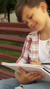 Neşeli beyaz bir çocuk canlı kırmızı bir bankta oturur, sıcak güneşin altında defterine neşeyle çizim yapar. Neşeli bir gülümseme ve dağınık esmer saçlarıyla, doğanın güzelliğini yakalar, çocukluğun saf neşesini somutlaştırır.