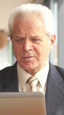 Özel dikim takım elbise ve kravat giymiş zarif bir kıdemli işadamı, şık bir şehir kafesinde laptopuna dikkatle odaklanırken dumanı tüten bir fincan kahveden zevk alıyor. Seçkin beyaz adam, çarpıcı gri saçları ve düşünceli bir ifadesi var.