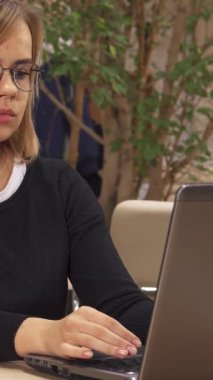 Genç beyaz bir kadın, gözle görülür şekilde stresli, yemyeşil ile dolu canlı bir çalışma alanında parlak bir masada otururken şakaklarına masaj yapıyor. Dikkatini dizüstü bilgisayara vermiş durumda. Baş ağrısıyla boğuşuyor, birden fazla serbest işle uğraşıyor.