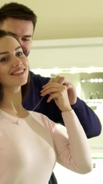 Zarif bir butikte, çekici sakallı bir adam sevgilisinin boynuna büyüleyici bir kolye takar ve onun neşeli gülümsemesini ön plana çıkarır. Güzel beyaz kadın saçlarını şakaya vurarak romantik bir ortam yaratıyor.