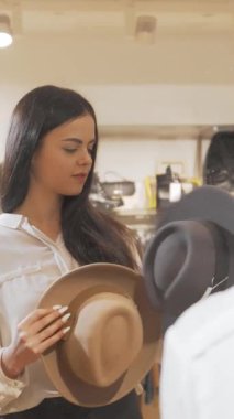 Çekici esmer bir kadın, parlak bir butikte şık kıyafetlerle çevrili aynanın önünde modaya uygun şapkalar denerken kendine güvenle ışıldıyor. Neşeli ifadesi ve şık seçimleri moda keşiflerinin heyecanını yansıtıyor.