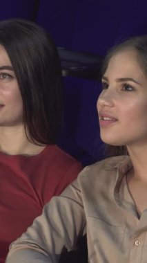 İki genç kadın, biri canlı kırmızı bluzlu, diğeri yumuşak bej bluzlu, samimi bir sinemada yan yana oturuyor, neşeli bir fısıltıyı paylaşırken yüzleri heyecandan parlıyor. Elinde patlamış mısır ve titreyen perdeyle