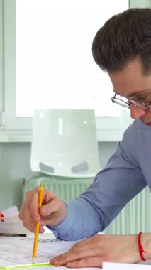 Gözlük takan kendini adamış genç beyaz bir mimar modern ofis masasında karmaşık bina planları çiziyor. Canlı planlar ve parlak turuncu bir başlıkla çevrelenmiş, profesyonellik ve yaratıcılık yayıyor, somutlaşıyor.