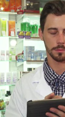 Gıcır gıcır beyaz bir tıbbi elbise giyen odaklanmış bir erkek eczacı, renkli ilaçlarla dolu canlı raflarla çevrili tabletindeki ilaç envanterini dikkatle inceliyor. Onun özenli ifadesi ve mesleki davranışı kritik durumun altını çiziyor.