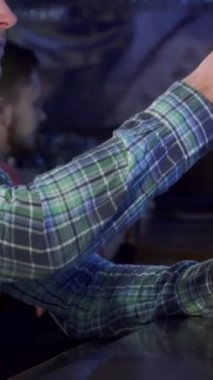 Tarz sahibi, kahverengi saçlı, beyaz bir adam soğuk birasından bir yudum alır, yan profili rahatlama ve keyif verir. Canlı bar atmosferi, kırsal ahşap barı ve sıcak, davetkar ışıklandırmasıyla, rahat bir zemin oluşturur.