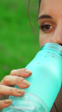 Neşeli genç bir kız şık spor şişesinden ferahlatıcı bir yudum alır, yoga çalışmasını canlı yeşil çimenlerin arasında durdurur. Işıl ışıl gülüşü ve gevşek tavırları, uyum ve doğayı yansıtıyor, sükunet hissi uyandırıyor.