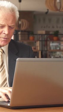 Özel dikim takım elbiseli ve çizgili kravatlı zarif, yaşlı beyaz bir adam laptopuna dalmış bir şekilde kırsal ahşap bir masada oturuyor ve dumanı tüten bir fincan kahvenin tadını çıkarıyor. Kafenin davetkar atmosferi, yumuşak ışıklandırma ve şıklıkla bezenmiş.