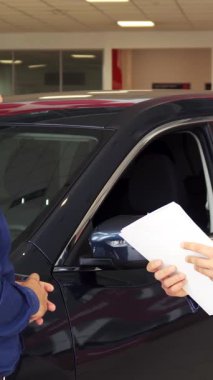 Modern bir araba galerisinde, şık giyimli bir satıcı ve memnun bir alıcı güven dolu bir tokalaşmayı paylaşıyor ve başarılı bir anlaşma imzalıyor. Hava heyecan ve profesyonellikle dolu, arka planda parıldayan parlak araç,