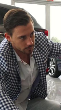 Koyu renk saçlı, bakımlı sakallı şık bir adam ışıldayan yeni arabasının yanında diz çöküp, ışıldayan bir gülümsemeyle baş parmağını kaldırıyor. Modaya uygun bir spor ceket ve kot pantolon giymişti. Parlak ve modern sergi salonunda kendine güveni tamdı.