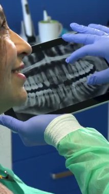 Kendini adamış genç bir beyaz dişçi, canlı yeşil koruyucu bir elbise giyer, tabletindeki detaylı x-ray görüntüsüne zum yapar, kadın bir hasta ise keskin bir ilgiyle gözlemler. Bu dinamik sahne modern diş bakımının sinerjisini vurgular.