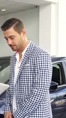 Modern bir araba galerisinde, şık giyimli bir adam iyi giyimli bir satıcıyla bir animasyon sohbeti yapar, her ikisi de bir belgede gösterilen etkileyici özelliklere odaklanır. Titreşimli atmosfer beklentiyle uğulduyor.