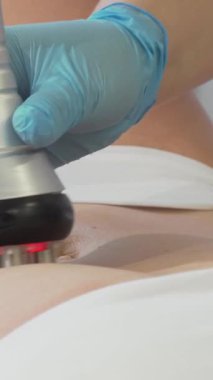 Kendini işine adamış bir güzellik uzmanı, bir kadın müşterinin midesine, dingin bir güzellik salonundaki ileri teknolojiyi vurgulamak için RF kaldırma cihazı uygular. Pürüzsüz, parlak cihaz kırmızı ışıklar yayıyor. Cildin sıkılaşmasını ve gençleşmesini sembolize ediyor.