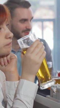 Kızıl saçlı genç bir kadın, arkadaşları yakınlardaki animasyon sohbetleriyle meşgulken hareketli bir barda tek başına oturmuş, buzlu bir bardak bira içiyor. Hüzünlü ifadesi, hayat dolu gece hayatının ortasında dokunaklı bir yalnızlık hissi yakalıyor.