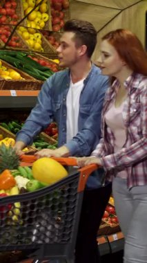 Neşeli genç bir çift, renkli bir ürün dizisiyle çevrili, hareketli bir alışveriş merkezinde taze sebze alışverişi yapıyor. Güzel kızıl saçlı kız, uzun dalgalı saçlarıyla alışveriş arabalarına gevrek bir lahana koyar.