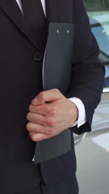 Özel dikim siyah takım elbiseli dengeli bir satış müdürü gururla sunar çarpıcı gri bir sedanın yanında bir araba anahtarı, profesyonellik ve başarı yayar. Düşük açılı dinamik perspektif hareketli bir bayiliğin canlı atmosferini yakalıyor.