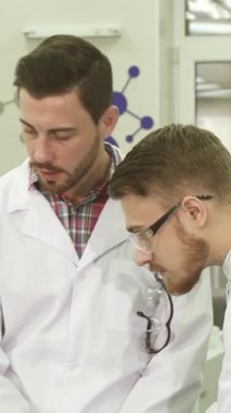 Parlak, modern bir laboratuvarda, canlı beyaz önlüklü ve mavi lateks eldivenli iki genç erkek asistan iş akışları hakkında animasyonlu bir tartışmaya girişirler. Odaklanmış ifadeleri ve çevrelerindeki parıldayan bilimsel ekipmanları