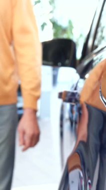 Neşeli, kıdemli, beyaz bir adam galerideki göz kamaştırıcı mor bir geçitin arka kapısını hevesle açıyor. Potansiyel yeni arabası için heyecanlanıyor. Yakın çekim, sergi salonunun ışıklarının parıldayan yansımalarını vurgular.