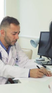 Parlak, modern bir muayenehanede şefkatli bir erkek doktor genç bir bayanla ciddi bir istişarede bulunurken röntgeni dikkatle inceler. Atmosfer profesyonellik ve özen gösteriyor.