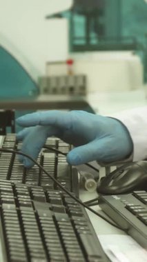 Kendini işine adamış beyaz önlüklü ve mavi lateks eldivenli bir erkek laboratuvar teknisyeni test tüplerinden düzgün bir bilgisayara ustalıkla veri giriyor. Bu canlı sahne teknoloji ve biyolojinin kesişimini vurgular, hassasiyeti somutlaştırır ve