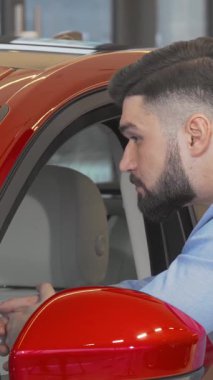 Şık gömlekli neşeli, sakallı bir adam galerideki göz kamaştırıcı kırmızı arabayı inceliyor, heyecan saçıyor. Kendine güveni ve çekici gülümsemesi mükemmel aracı keşfetme heyecanını yakalıyor, modern tüketiciliği somutlaştırıyor.