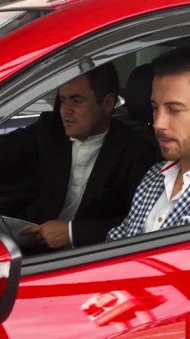 Çarpıcı kırmızı bir arabanın içinde, şık giyimli bir satıcı ve özenli bir alıcı modern iç mekanı eşeliyor, yüksek teknoloji gösterge panelini ve kontrolleri inceliyor. Canlı gösteri salonu atmosferi, lüks arabalar hakkındaki ilgi çekici tartışmalarını güçlendiriyor.