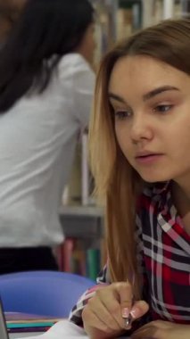 Dalgalı kahverengi saçlı kendini adamış genç bir kadın notlarını dikkatle not defterine yazar. Çalışmalarına canlı bir kütüphanede dalmıştır. Arka planda, kitaplıklarla ilgilenen bir çift, akademik ortama hayat dolu bir ortam ekliyor.